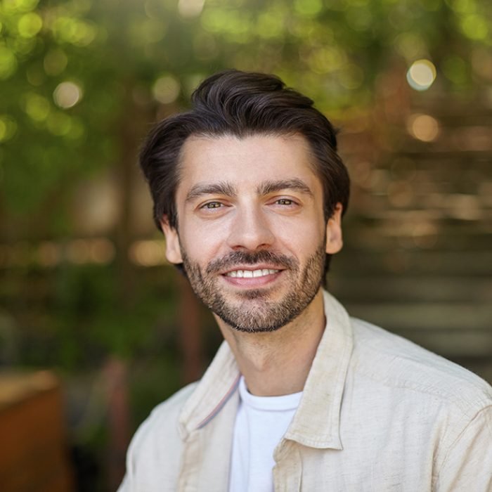 Close-up of beautiful bearded male with dark hair posing over green ciy park on sunny day, looking to camera with charming and sincere smile