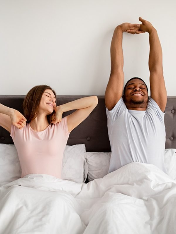 Young multiracial couple stretching on bed after waking up at home. Millennial family having lazy weekend morning, enjoying cozy times together, feeling sleepy, relaxing on Sunday indoors